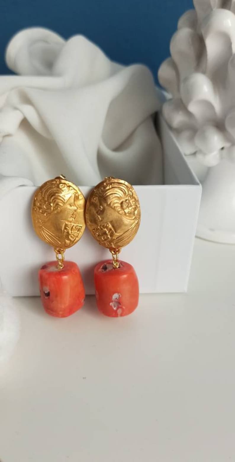 Pu&ograve; includere: Orecchini dorati con un designo floreale dettagliato e perline color corallo che pendono dalla parte inferiore.