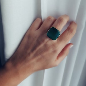 Könnte beinhalten: Ein quadratischer Ring mit einem dunkelgrünen Edelstein, der in eine goldfarbene Metallfassung gefasst ist.
