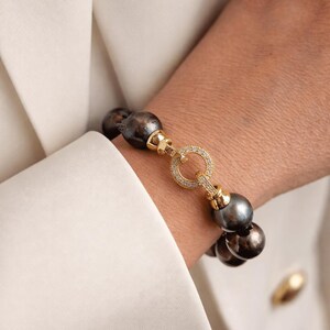 May include: A bracelet with dark grey pearls and gold-coloured metal detailing. The bracelet has a circular clasp set with small, clear gemstones. The background shows a white blazer. The pearls are approximately 1 cm in diameter.