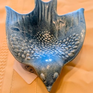 Könnte beinhalten: Eine blaue Keramikschale in Vogelform mit strukturierter Oberfläche. Die Schale hat einen spitzen Schnabel und ausgebreitete Flügel. Die Flügel und der Kopf haben eine glatte, glänzende Oberfläche, während der Körper ein gepunktetes Muster aufweist.