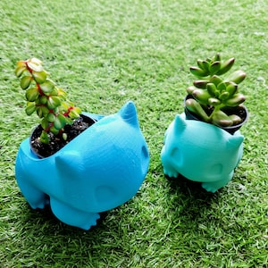 Puede incluir: Dos macetas azules con forma de personaje de dibujos animados, cada una con una pequeña planta suculenta. La maceta más grande es de color azul brillante, mientras que la más pequeña es de color turquesa claro. Las plantas tienen hojas verdes sobre un fondo verde.