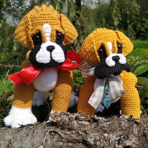 Pode incluir: Dois peluches de cachorro boxer de crochê, um com um laço vermelho e o outro com uma fita branca e cinza, sentados em um galho de árvore.