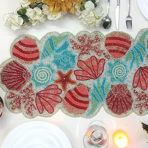 May include: A beaded table runner with a sea life design. The runner features red, teal, and white seashells, starfish, and coral. The runner is on a white surface, with a candle, flowers, and silverware visible.