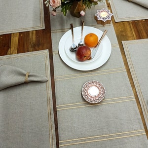 Op de afbeelding: Een set van vier grijze linnen placemats met een gouden rand. De placemats liggen op een houten tafel met een wit bord, bestek, fruit en een kaarsenhouder.