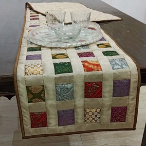 Könnte beinhalten: Ein beiger Tischläufer mit braunem Rand. Der Läufer besteht aus Stoffquadraten in verschiedenen Farben und Mustern, darunter Grün, Lila, Rot und Gold.