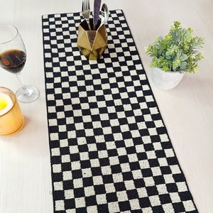 Puede incluir: Un camino de mesa a cuadros blanco y negro hecho de cuentas. El camino de mesa está sobre una mesa blanca con un jarrón de metal dorado y una copa de vino tinto.