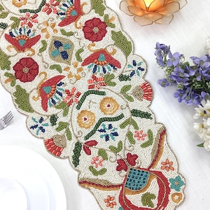 Puede incluir: Un camino de mesa largo con bordes festoneados y abalorios intrincados sobre una base color crema. El diseño muestra un patrón floral vibrante con cuentas de vidrio rojas, verdes, azules y naranjas dispuestas en motivos botánicos detallados.