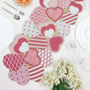 May include: A beaded heart-shaped table runner in shades of pink, white, and red. The runner features various patterns, including stripes, diamonds, and solid hearts. It is designed for a festive table setting, perfect for Valentine's Day or a romantic dinner.