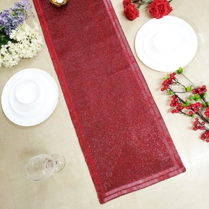 May include: A red beaded table runner with a subtle shimmer. The runner is placed on a light wood surface with white plates, a glass, and decorative flowers.