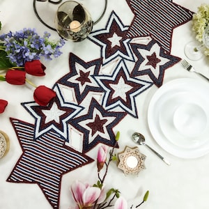 May include: A red, white, and blue beaded table runner with a star design. The runner is made of small beads and has a striped pattern. The runner is on a white tablecloth with candles and flowers.