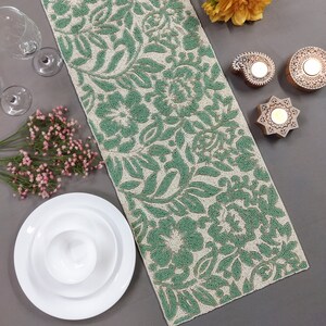 May include: A green and white beaded table runner with a floral pattern. The runner is on a gray surface with white plates and flowers.