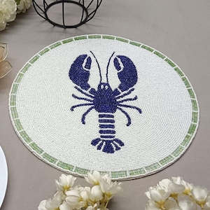 Peut inclure: Un dessous de plat rond, orné d'un motif de homard bleu marine sur fond blanc perlé. Le bord est bordé d'une bordure verte. Il est posé sur une surface grise, avec des fleurs et un bougeoir en arrière-plan.
