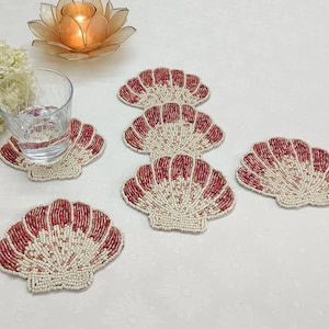 May include: Five red and white beaded seashell-shaped coasters. The coasters are arranged in a semi-circle on a white surface.