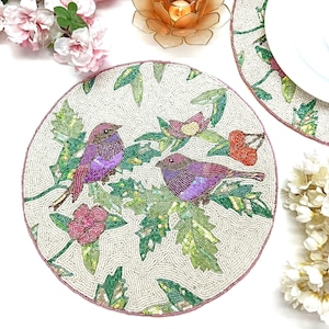 May include: A set of two round beaded placemats with a white background and a floral design featuring pink flowers, green leaves, and two purple birds.