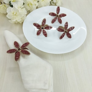 May include: Three burgundy beaded flower napkin rings on a white plate and one on a white napkin. The napkin rings are made of gold wire and have a beaded flower design.