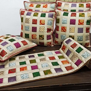 K&ouml;nnte beinhalten: Ein Set aus f&uuml;nf cremefarbenen Kissen mit Patchwork-Muster, das aus Quadraten in verschiedenen Farben besteht, darunter Rot, Gr&uuml;n, Lila und Gold. Die Kissen sind auf einem Tischl&auml;ufer mit &auml;hnlichem Patchwork-Muster angeordnet.