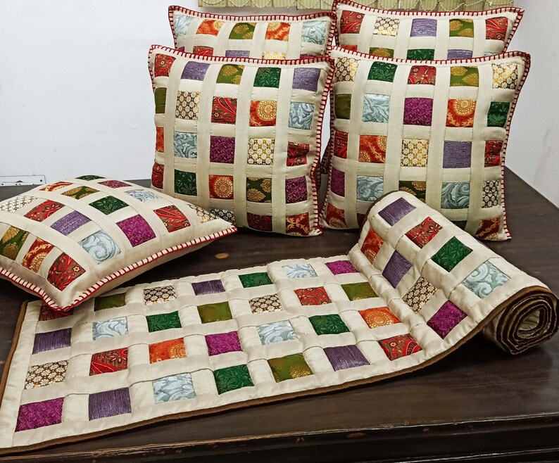 K&ouml;nnte beinhalten: Ein Set aus f&uuml;nf cremefarbenen Kissen mit Patchwork-Muster, das aus Quadraten in verschiedenen Farben besteht, darunter Rot, Gr&uuml;n, Lila und Gold. Die Kissen sind auf einem Tischl&auml;ufer mit &auml;hnlichem Patchwork-Muster angeordnet.