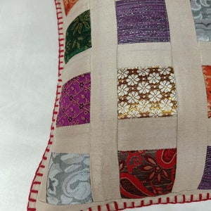 K&ouml;nnte beinhalten: Ein beigefarbenes Kissen mit Patchwork-Muster aus Quadraten aus verschiedenen bunten Stoffen. Das Kissen hat einen roten und wei&szlig;en gestickten Rand.
