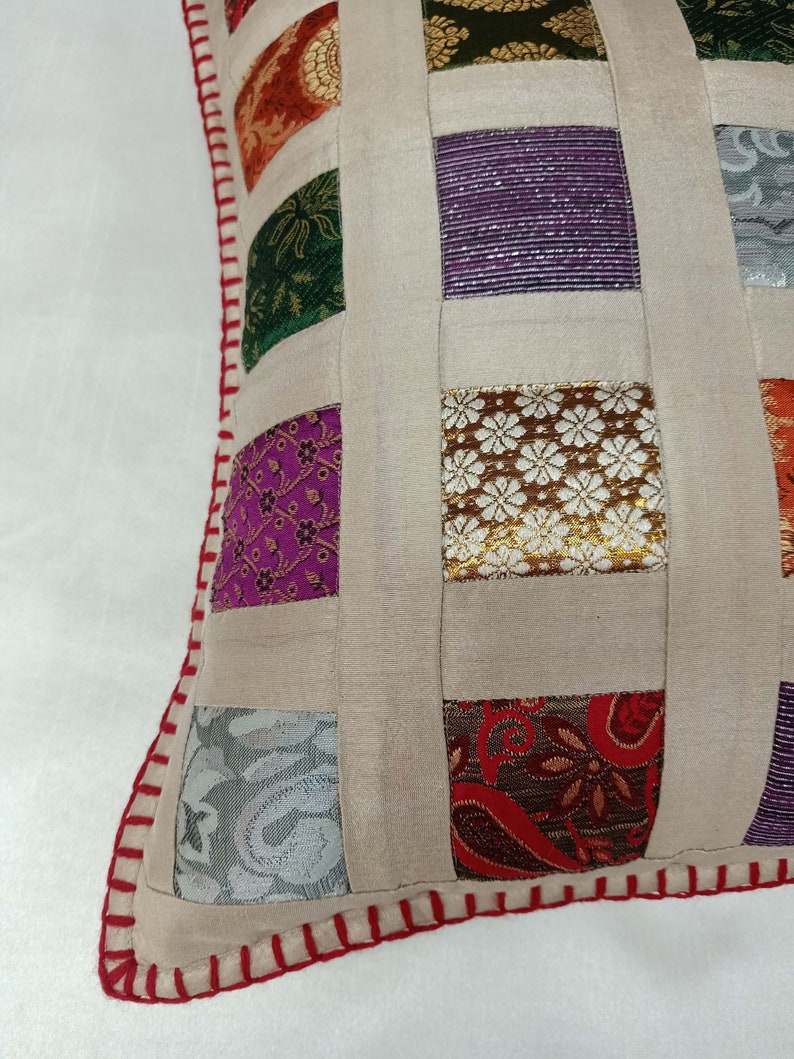 K&ouml;nnte beinhalten: Ein beigefarbenes Kissen mit Patchwork-Muster aus Quadraten aus verschiedenen bunten Stoffen. Das Kissen hat einen roten und wei&szlig;en gestickten Rand.