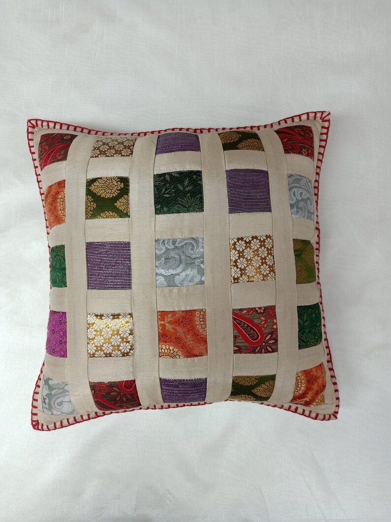 K&ouml;nnte beinhalten: Ein quadratisches Kissen mit Patchwork-Muster. Das Kissen ist aus beigefarbenem Stoff mit Quadraten in verschiedenen Farben und Mustern, darunter Lila, Gr&uuml;n, Gold und Rot. Das Kissen hat einen roten und wei&szlig;en gestickten Rand.