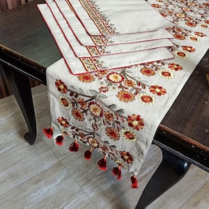 Op de afbeelding: Een set van vier geborduurde placemats en een bijpassende tafelrunner met een bloemenmotief in tinten rood, oranje en geel. De placemats en runner hebben een crèmekleurige achtergrond en een rode rand.