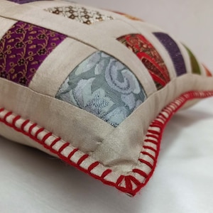 K&ouml;nnte beinhalten: Ein cremefarbenes Kissen mit einem roten, gesteppten Rand. Das Kissen besteht aus Patchwork-Stoff mit verschiedenen Mustern und Farben, darunter Lila, Gr&uuml;n und Blau.