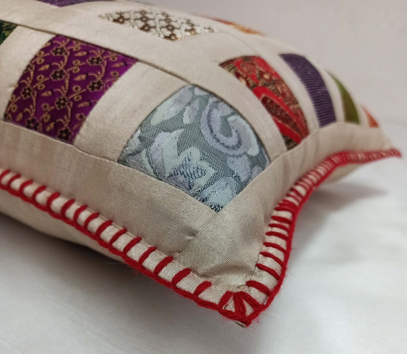 K&ouml;nnte beinhalten: Ein cremefarbenes Kissen mit einem roten, gesteppten Rand. Das Kissen besteht aus Patchwork-Stoff mit verschiedenen Mustern und Farben, darunter Lila, Gr&uuml;n und Blau.