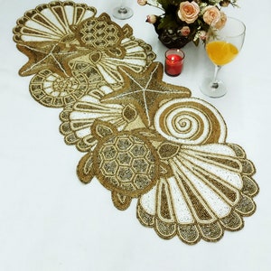 May include: A beaded table runner with a seashell, starfish, and turtle design. The runner is made of white and gold beads and is perfect for a coastal-themed table setting.