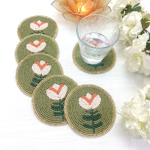 May include: A set of beaded coasters with a floral design. The coasters are round, with a green background and a white and orange flower in the center. The edges are trimmed with gold beads. A glass of water sits on one coaster.