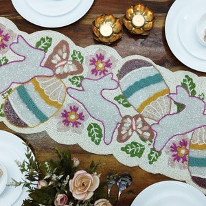 May include: A white beaded table runner with a pastel Easter theme. The runner features butterflies, bunnies, and eggs with a multi-coloured striped design.