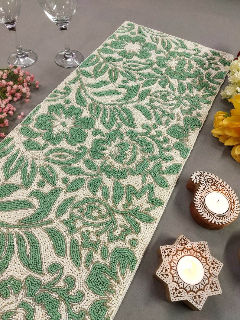 May include: A green and white beaded table runner with a floral design. The runner is made of small beads that are sewn together to create a intricate pattern. The runner is perfect for adding a touch of elegance to any table setting.