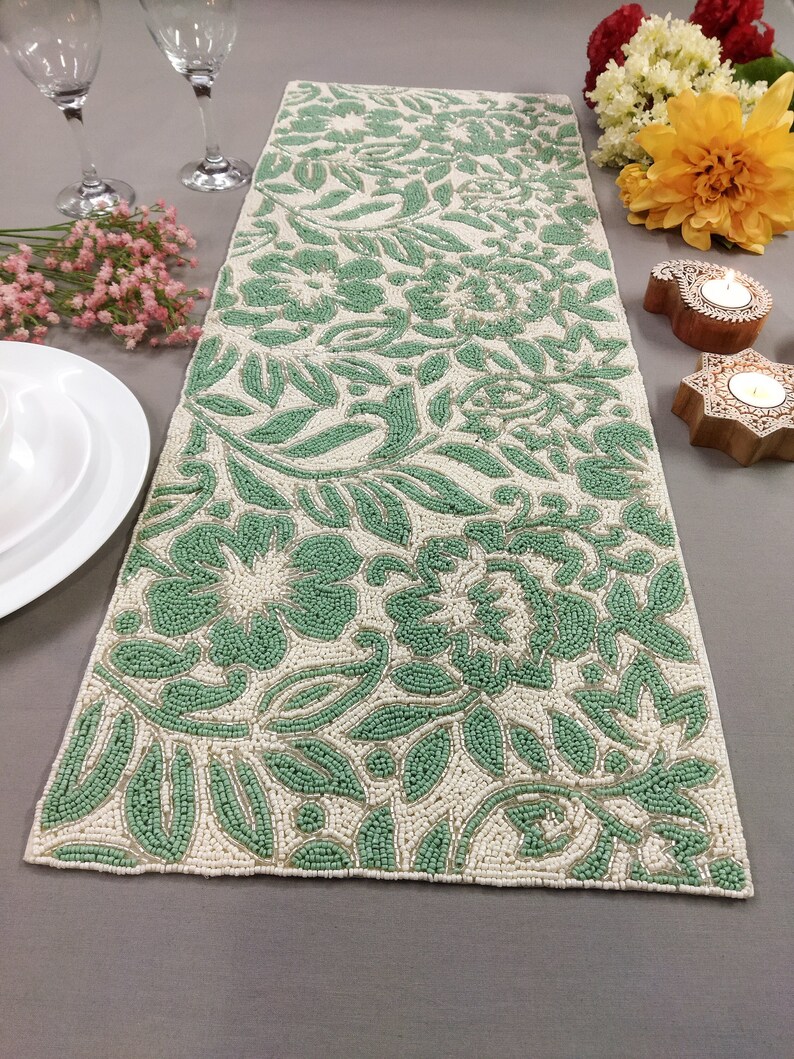 May include: A green and white beaded table runner with a floral pattern. The runner is made of small beads that are sewn together to create a raised design.