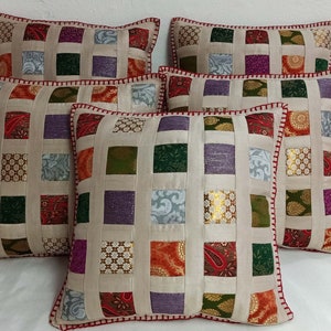 K&ouml;nnte beinhalten: F&uuml;nf dekorative Kissen mit Patchwork-Muster. Die Kissen sind aus beigefarbenem Stoff mit Quadraten in verschiedenen Farben und Mustern, darunter Gr&uuml;n, Lila, Rot und Gold. Die Kissen haben einen roten und wei&szlig;en Rand.
