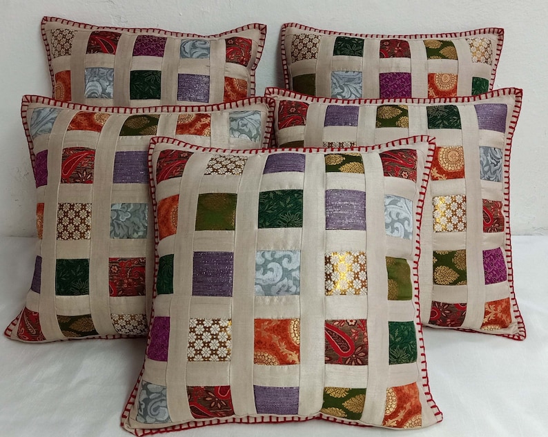 K&ouml;nnte beinhalten: F&uuml;nf dekorative Kissen mit Patchwork-Muster. Die Kissen sind aus beigefarbenem Stoff mit Quadraten in verschiedenen Farben und Mustern, darunter Gr&uuml;n, Lila, Rot und Gold. Die Kissen haben einen roten und wei&szlig;en Rand.
