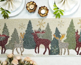 May include: A beaded table runner with a winter forest scene. The runner features a beige background with green trees, white snowflakes, and red and brown reindeer.