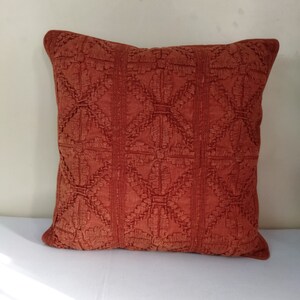 May include: A square, rust-colored decorative pillow with an embroidered geometric pattern. The pillow features a repeating design of diamond shapes and vertical stripes, adding texture and visual interest. The pillow is a warm, inviting accent for any living space.