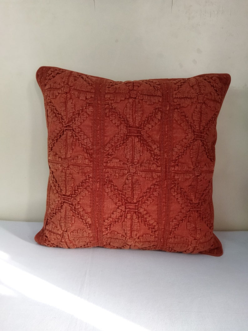 May include: A square, rust-colored decorative pillow with an embroidered geometric pattern. The pillow features a repeating design of diamond shapes and vertical stripes, adding texture and visual interest. The pillow is a warm, inviting accent for any living space.