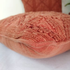 May include: Close-up of two terracotta-colored decorative pillows. The front pillow has a textured, fringed design, while the pillow in the background has a diamond pattern. The pillows are on a white surface.