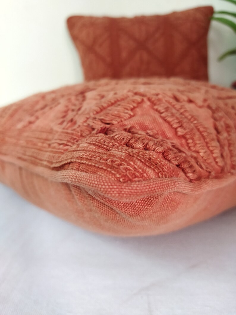 May include: Close-up of two terracotta-colored decorative pillows. The front pillow has a textured, fringed design, while the pillow in the background has a diamond pattern. The pillows are on a white surface.