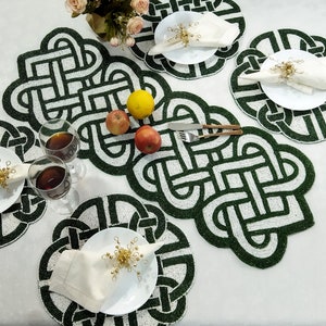 May include: A table setting featuring green and white beaded placemats and a matching table runner. White plates hold folded napkins secured by gold napkin rings. Wine glasses and fruit complete the elegant dining arrangement.