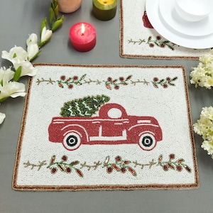 Puede incluir: Salvamanteles de cuentas blancas con un camión rojo que lleva un árbol de Navidad verde. El camión tiene ruedas negras y un fondo blanco. El salvamanteles tiene un borde dorado y un diseño de hojas verdes y bayas rojas.