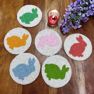 May include: Six round beaded coasters featuring different coloured bunny silhouettes. The coasters are white with green, orange, pink, red, blue, and green bunnies.