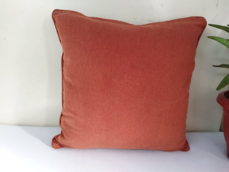 May include: A square, terracotta-colored pillow with a soft, textured surface. The pillow has a simple design with a stitched border. The pillow is placed on a white surface, with a plant in a red pot visible in the background.