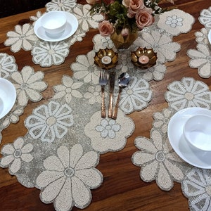 Pode incluir: Um conjunto de seis tapetes de mesa de contas brancas com um design floral. Os tapetes de mesa estão dispostos em uma mesa de madeira com tigelas e talheres.