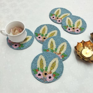 May include: Set of five blue beaded coasters with a bunny rabbit design. Each coaster features a pair of bunny ears, a floral pattern, and a light blue background.