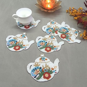 May include: White beaded teapot-shaped coasters with floral designs in blue, orange, and green. A white teacup sits on one coaster. The coasters are arranged on a gray surface, with a candle and decorative flowers in the background.