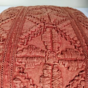 May include: Close-up of a terracotta-colored decorative pillow. The pillow features an intricate, raised embroidery pattern with a geometric and floral design. The edges are finished with a detailed, textured border.