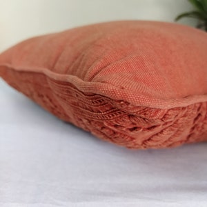 May include: A terracotta-colored decorative pillow with a textured, woven fabric. The pillow has a detailed, embroidered border. The pillow is on a white surface. Home decor item.