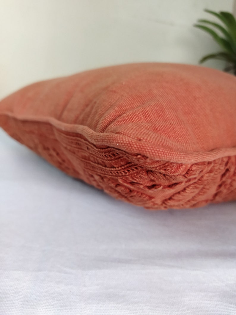 May include: A terracotta-colored decorative pillow with a textured, woven fabric. The pillow has a detailed, embroidered border. The pillow is on a white surface. Home decor item.