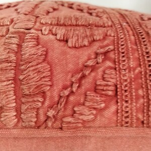 May include: Close-up of a coral-colored decorative pillow. The pillow features a textured design with raised, fringed elements and a detailed border. The fabric appears to be a woven material, adding to the tactile quality of the pillow.